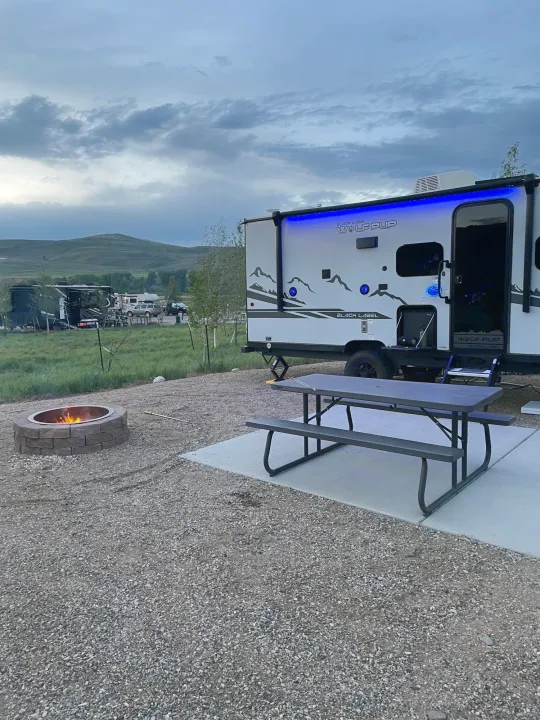 RV rental campsite: A white travel trailer with blue lights, a picnic table, and a burning fire pit. Cloudy sky, hills.. Forest River Cherokee Wolf Pup Black Label 2022