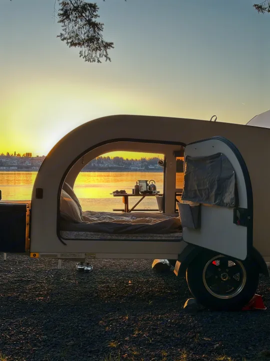 An open RV rental teardrop camper sits waterfront at sunset, revealing a cozy bed and compact kitchen inside.. DROPLET XL 2024