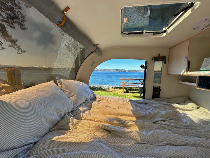 Relax in your RV rental's comfy bed. Enjoy a waterfront view of a lake, picnic table, and distant city from inside.. DROPLET XL 2024
