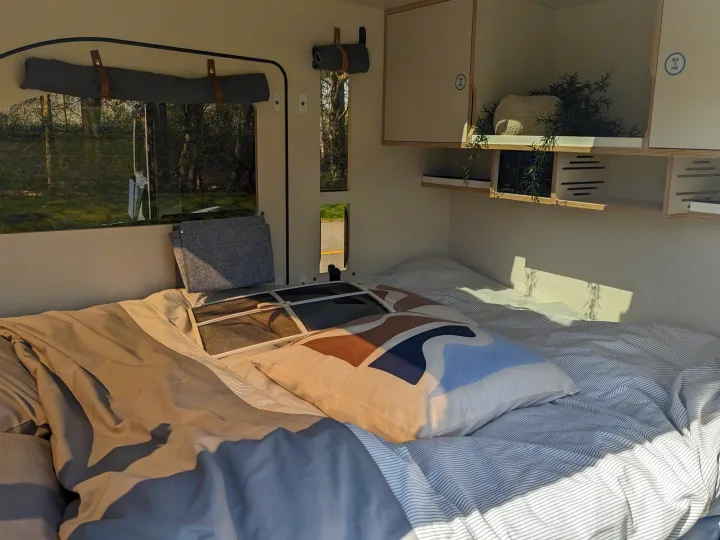 Cozy RV rental bedroom with a comfortable bed, striped bedding, compact storage, and scenic window views of nature.. DROPLET XL 2024