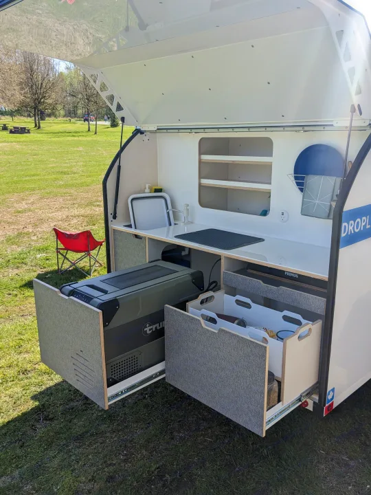An open teardrop trailer's outdoor kitchen, featuring a pull-out fridge, sink, and storage, ready for your next RV rental adventure.. DROPLET XL 2024