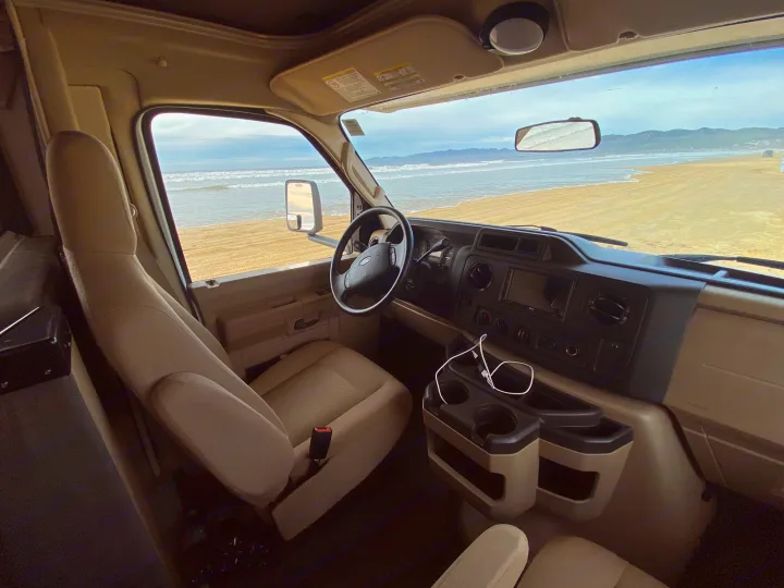 Inside an RV rental, the beige driver's cabin overlooks a sandy beach and ocean. Ideal for adventurous road trips.. Coachmen Leprechaun 2021