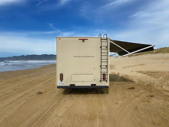 An RV rental on a sandy beach. This beige camper, with its awning extended, faces the ocean, offering seaside outdoor adventure.. Coachmen Leprechaun 2021