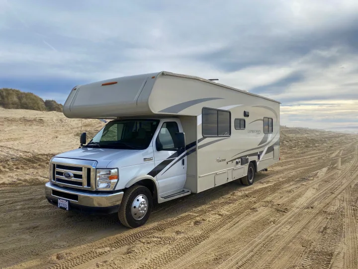 White Class C RV rental parked on a vast sandy beach under a cloudy sky, ready for scenic outdoor adventures.. Coachmen Leprechaun 2021