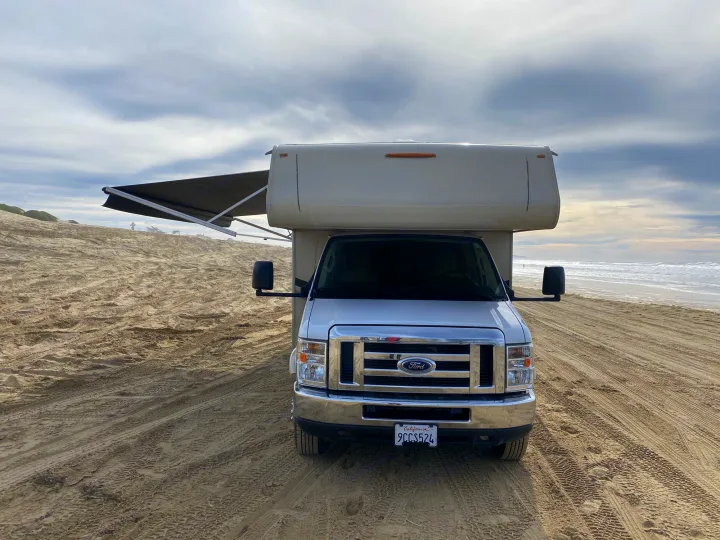 Front view of a white Ford RV rental with its awning out, parked on a wide sandy beach. The ocean and a cloudy sky are in the background.. Coachmen Leprechaun 2021