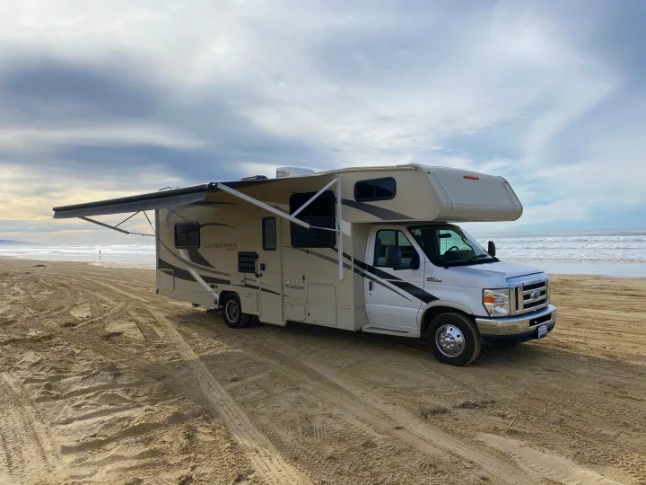 RV rental parked directly on a vast sandy beach by the ocean, awning extended. Explore coastal travel & adventure.. Coachmen Leprechaun 2021