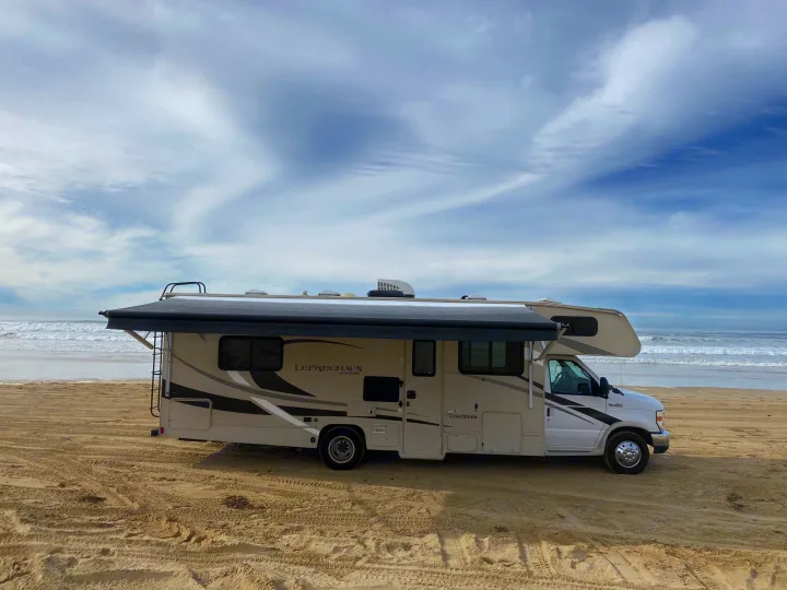 An RV rental is parked on a sandy beach by the ocean, with its awning extended. Enjoy coastal adventures with this Class C RV.. Coachmen Leprechaun 2021