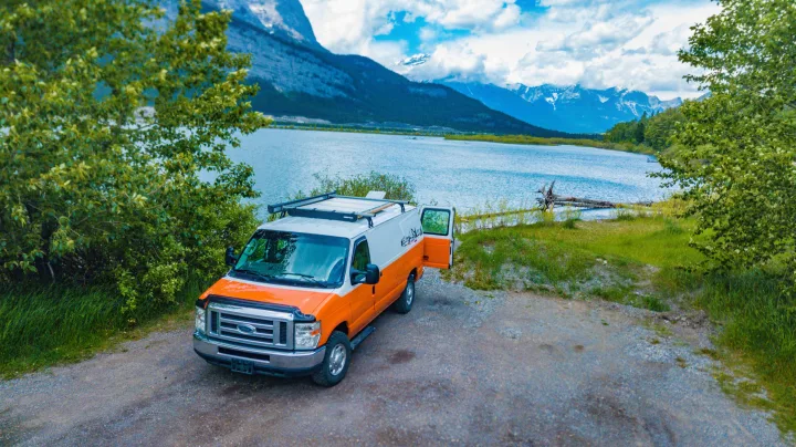 Your next **rv rental**: an orange and white campervan by a serene mountain lake, mountains in the background. Perfect for outdoor adventures!. Ford Econoline E250supercab 2012