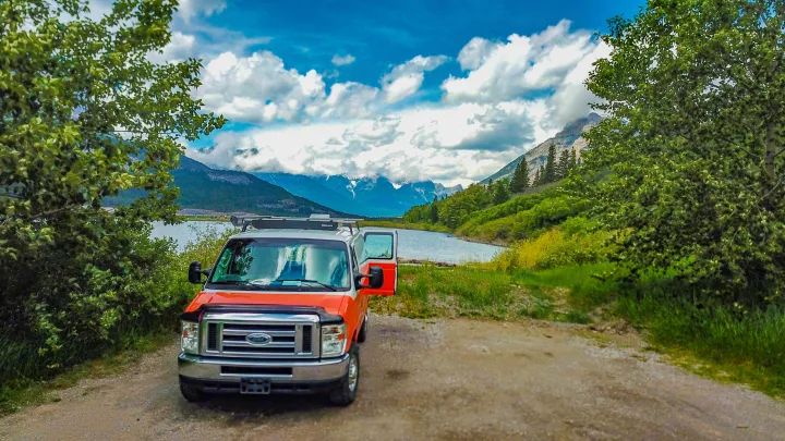 Orange & white RV rental van parked by a scenic lake with mountains under a cloudy sky. Perfect for a nature road trip.. Ford Econoline E250supercab 2012