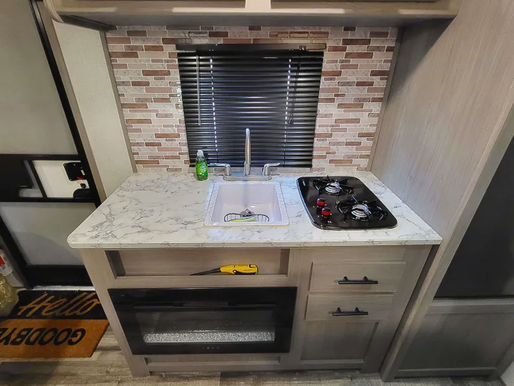 Compact RV rental kitchen features a sink, two-burner stove, marble-pattern counter, and stylish tiled backsplash.. Dutchmen Coleman Lantern LT 2023