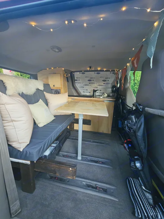 Inside a comfy RV rental van: a bench with cushions, portable table, wood cabinetry, and string lights create a cozy mobile home.. Dodge Caravan 2003