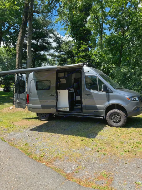 Modern gray Mercedes Sprinter RV rental with open side showing interior, parked under trees with awning extended.. Winnebago Revel 2023