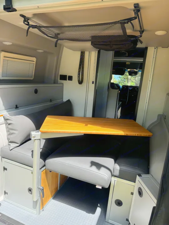 This RV rental features a compact interior dining area with a bamboo table, gray cushioned benches, and overhead storage net.. Winnebago Revel 2023
