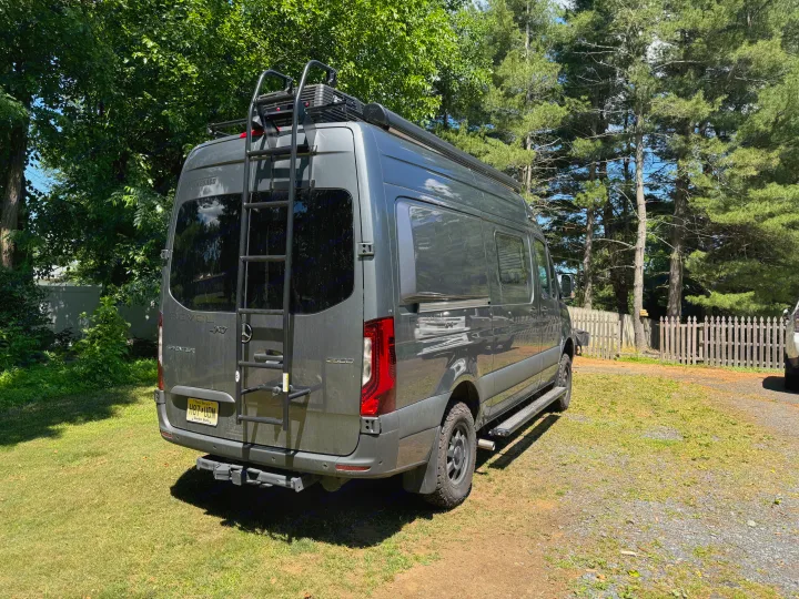A gray Winnebago Revel Sprinter 4x4 RV with roof rack and ladder, perfect for rv rental trips, parked outdoors.. Winnebago Revel 2023