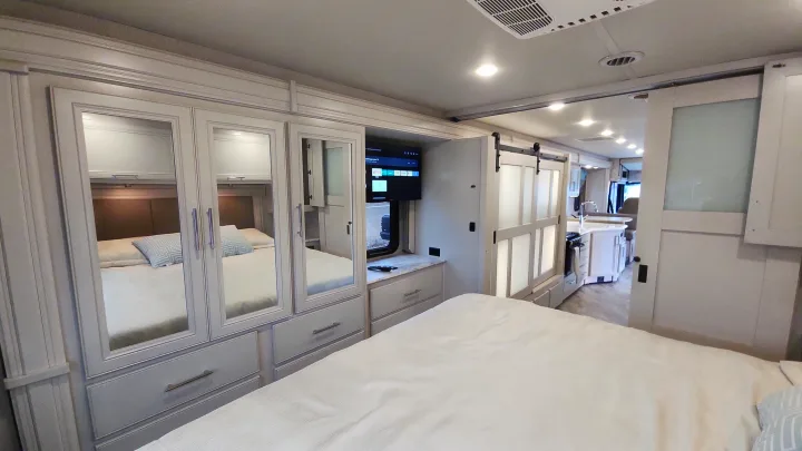 Modern RV rental interior: bedroom with mirrored closets, TV, and view of the kitchen through a sliding barn door.. Holiday Rambler Eclipse 2024