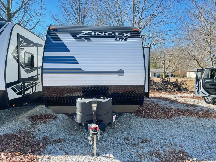Front view of a white Zinger Lite rv rental trailer on gravel. Bare trees & a truck are visible in the sunny outdoor setting.. Crossroads RV Zinger Lite 2021
