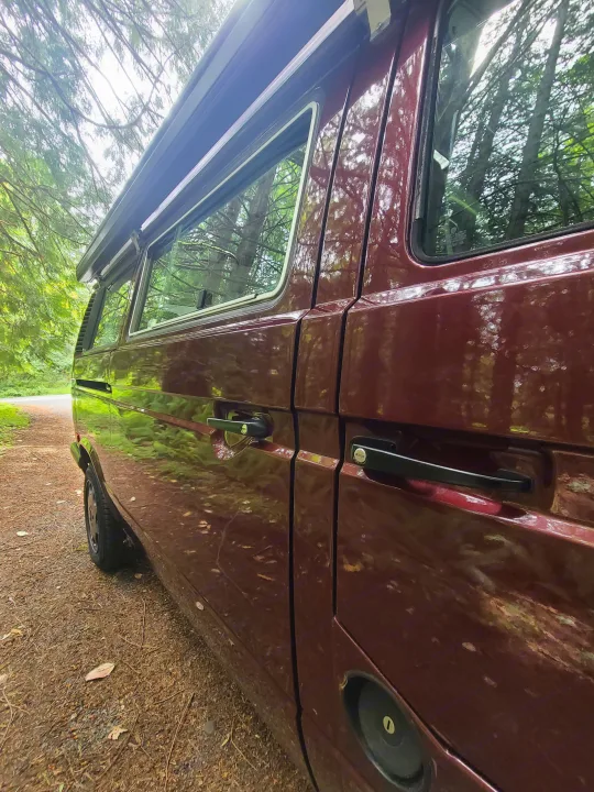 This burgundy camper van, an RV rental, is parked on a forest road. Its shiny paint reflects trees, with an awning above.. Volkswagen T3 Mulitvan 1991