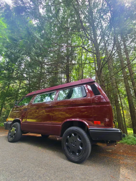 Low-angle view of a maroon pop-top VW camper van parked on an asphalt road in a lush forest, perfect for an RV rental adventure.. Volkswagen T3 Mulitvan 1991
