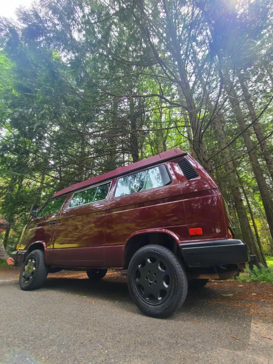 A deep red VW Vanagon camper van, an RV rental, parked on a forest road with sunlight filtering through tall trees.. Volkswagen T3 Mulitvan 1991