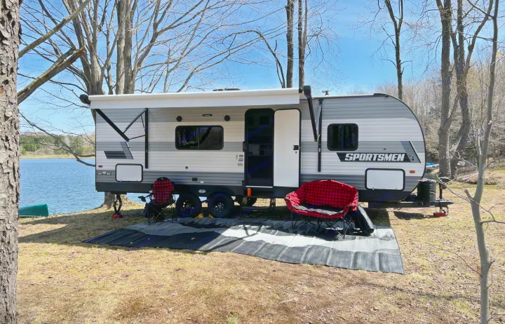 Experience this scenic RV rental: a travel trailer parked lakeside, complete with awning, outdoor chairs, and canoe for nature escapes.. KZ Sportsmen 26BHSE 2022