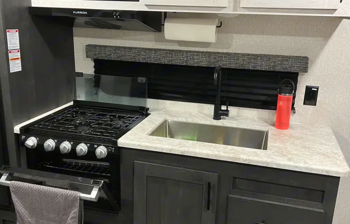 Inside an RV rental: a compact kitchen with a gas stove, oven, sink, and stylish dark cabinets.. KZ Sportsmen 26BHSE 2022