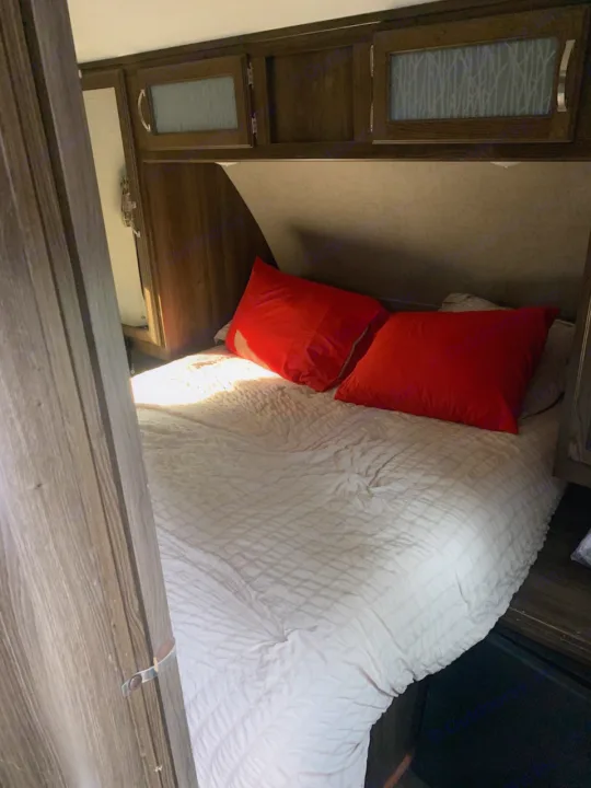 Inside an RV rental: a cozy bedroom features a bed with white comforter, bright red pillows, and overhead storage cabinets.. Keystone RV Bullet Ultra Lite 2018