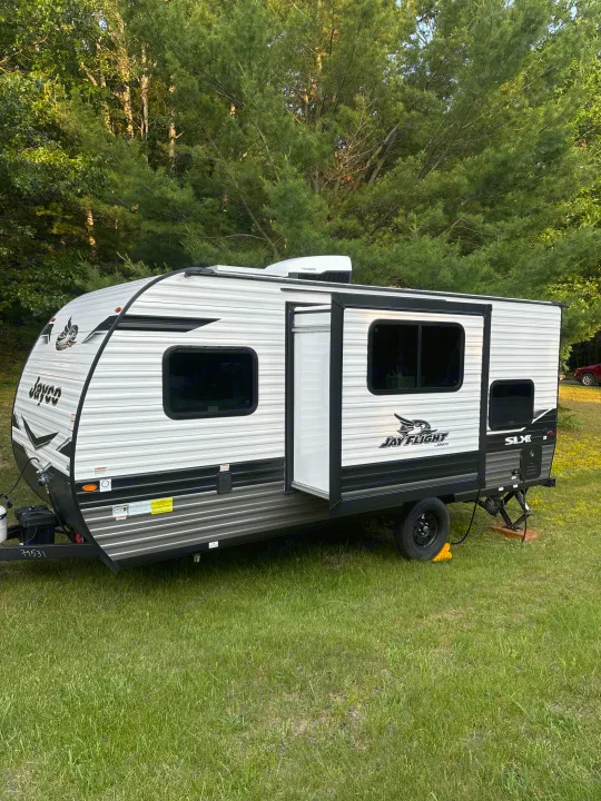 A white and black Jayco RV rental sits on green grass amidst tall trees, door open, ready for outdoor adventure.. Jayco Jay Flight SLX 2024