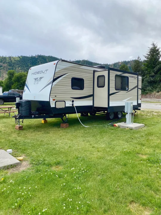 A Hideout travel trailer, an RV rental, is set up on grass at a scenic mountain campground with utilities connected.. Keystone RV Hideout 2018