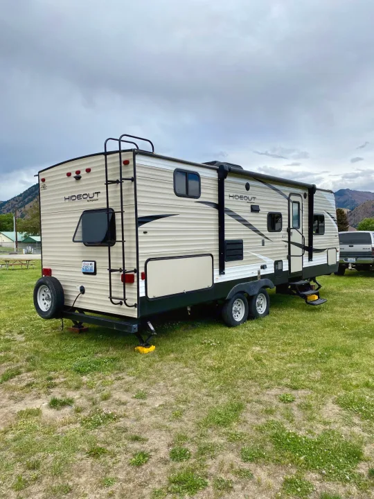 This beige "Hideout" RV rental travel trailer with extended steps is parked on grass under a cloudy sky, ready for adventure.. Keystone RV Hideout 2018