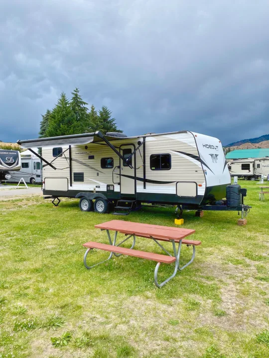 A beige "Hideout" rv rental is parked on a grassy campsite with an open awning & red picnic table under a cloudy sky.. Keystone RV Hideout 2018