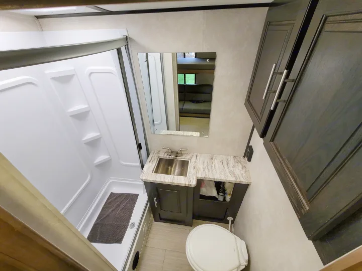 Compact RV rental bathroom featuring a shower, sink vanity, toilet, and overhead cabinets.. Forest River FR3 2020