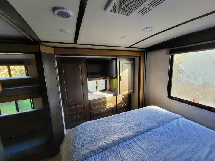 Cozy rv rental bedroom showing a queen bed, integrated storage, TV, and sunlight from windows. Bunk beds are visible on the left.. Forest River FR3 2020