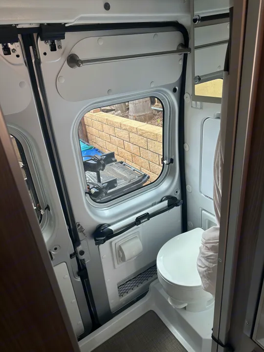 Compact RV rental camper van bathroom with a toilet, window, and wall-mounted storage including a towel bar.. Thor Sequnce 2023