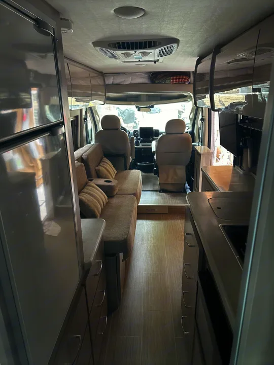 Bright RV rental interior, showing the living area with sofa, kitchen with counter, fridge, TV, and front cockpit. Overhead bunk visible.. Thor Sequnce 2023