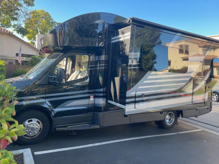 Sleek black & grey Class C RV rental, a Mercedes-Benz Sprinter chassis, parked on asphalt with houses and trees in the background.. Thor Motor Coach Delano 2022