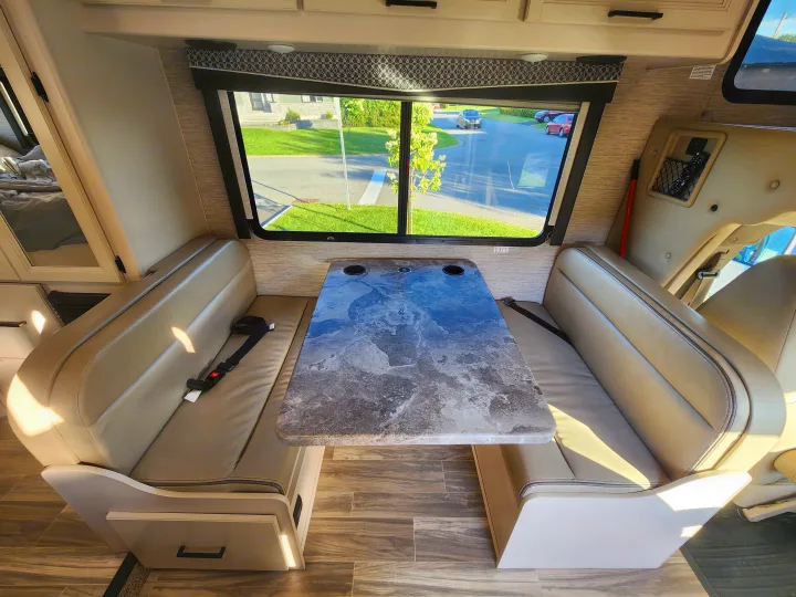 RV rental interior featuring a cozy dinette with tan seating, a marbled table, and large windows revealing the outdoors.. Thor Four Winds 2024