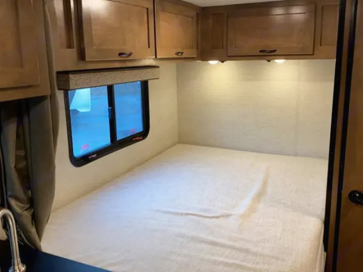 Inside an RV rental: A comfy bed with light sheets, overhead cabinets, and a window. Under-cabinet lights illuminate the wall.. Tiffin Motorhomes Wayfarer 2021