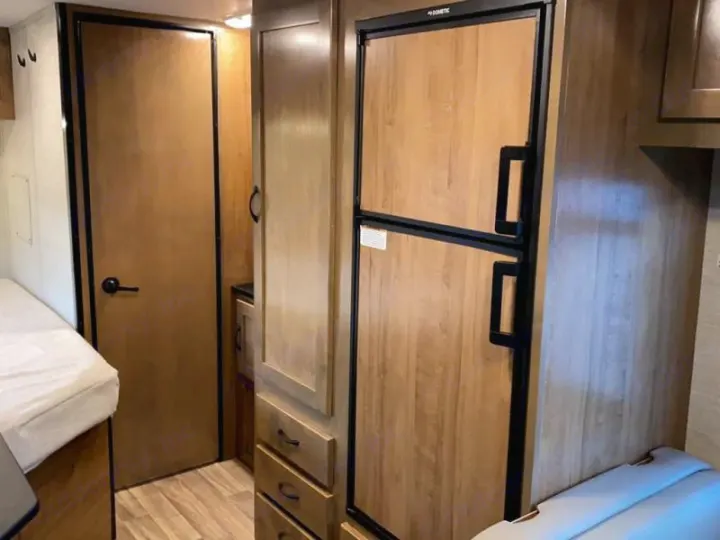 Cozy RV rental interior: visible bed, wooden door, storage cabinet, and a built-in refrigerator. Perfect for your next adventure.. Tiffin Motorhomes Wayfarer 2021