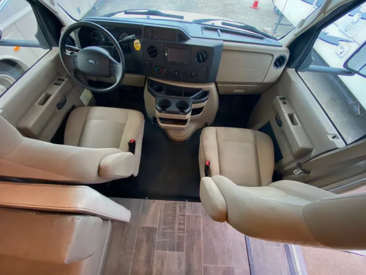 Inside an RV rental, showcasing the cockpit with beige seats, Ford steering wheel, dashboard, and center console with cup holders.. Winnebago 25J 2019