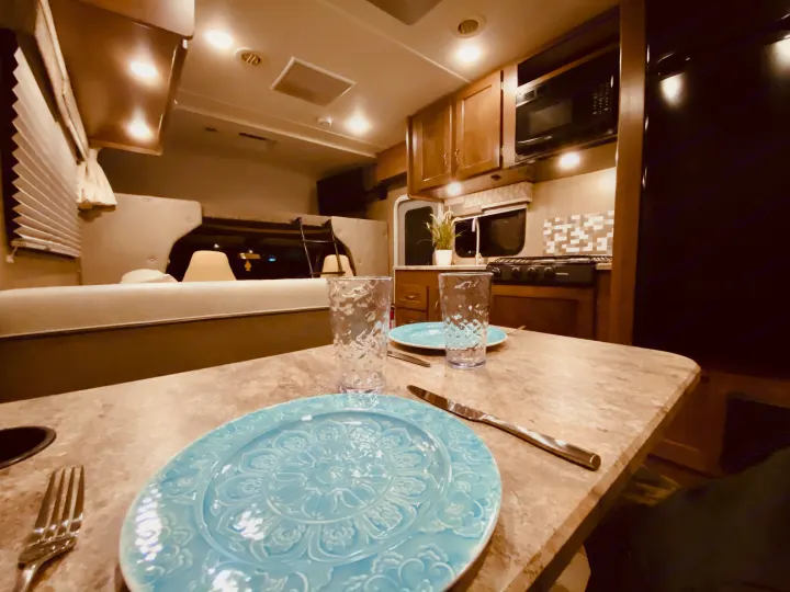 Cozy RV rental interior with a dining table set for two, a compact kitchen, and a sleeping alcove in the background.. Winnebago 25J 2019