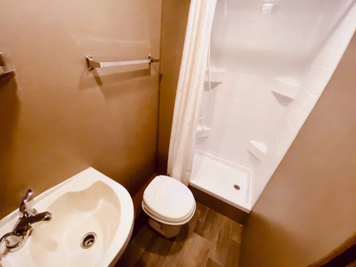 Interior view of an RV rental bathroom, showing a space-saving sink, toilet, and shower stall with a white curtain.. Winnebago 25J 2019