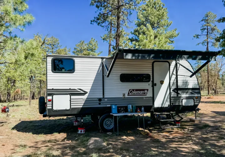 An RV rental, a white Coleman travel trailer, set up at a sunny forest campsite with an awning and a red generator visible.. Dutchmen Lantern LT BB19 2024