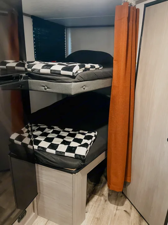 Upper and lower bunk beds with checkered blankets in an RV rental, featuring a privacy curtain.. Dutchmen Lantern LT BB19 2024