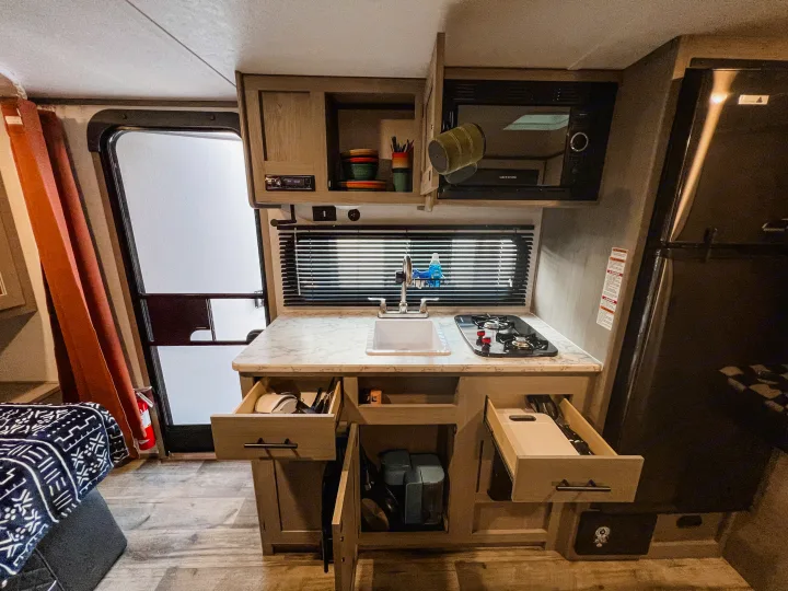 Explore this rv rental's compact kitchen, showcasing a sink, stovetop, microwave, and organized open drawers.. Dutchmen Lantern LT BB19 2024