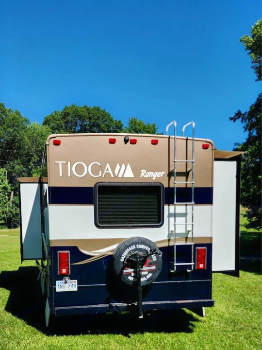 Rear view of a Tioga Ranger rv rental, tan & blue, parked on grass. Features include a ladder, spare tire, and window under a clear sky.. Tioga Ranger 2013