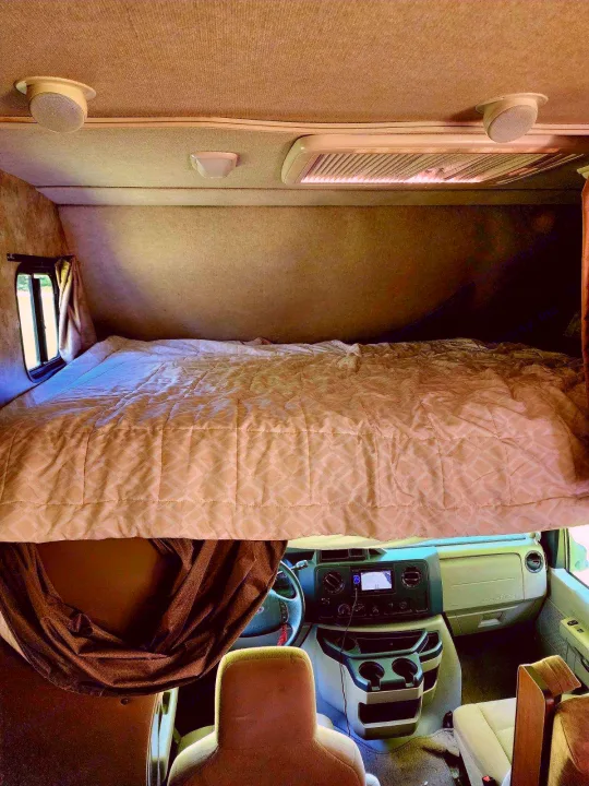 Cozy over-cab bed with a quilted comforter in this RV rental, overlooking the driver's cockpit and dashboard.. Tioga Ranger 2013