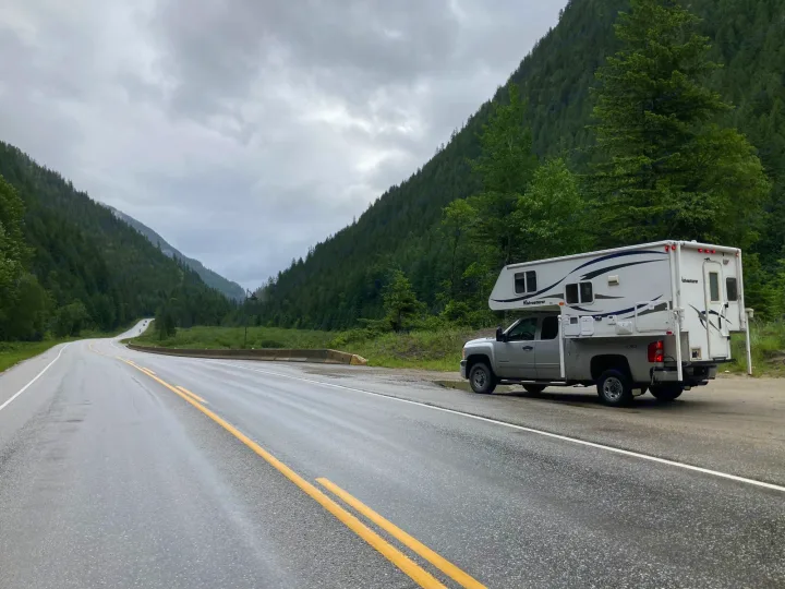 Scenic mountain road trip with an RV rental. A truck camper is parked on a wet highway shoulder, surrounded by lush green forests and grey skies.. Chevrolet Other 2010