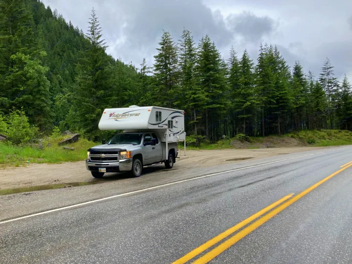 Silver pickup with "Adventurer" truck camper parked on a gravel shoulder next to a wet road and dense forest. Ideal for an RV rental.. Chevrolet Other 2010