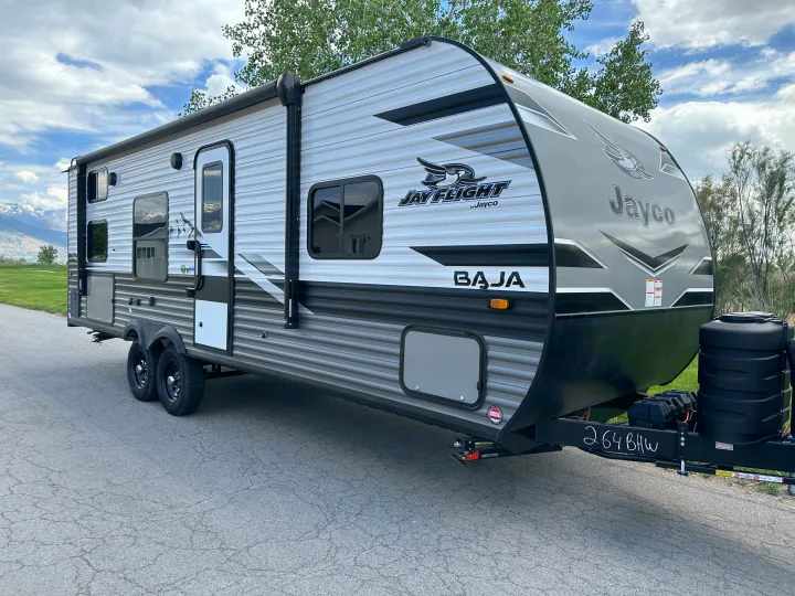 Modern gray Jayco Jay Flight Baja travel trailer RV rental, parked on asphalt with scenic mountains and green fields.. Jayco Jay Flight SLX Baja Edition 2023