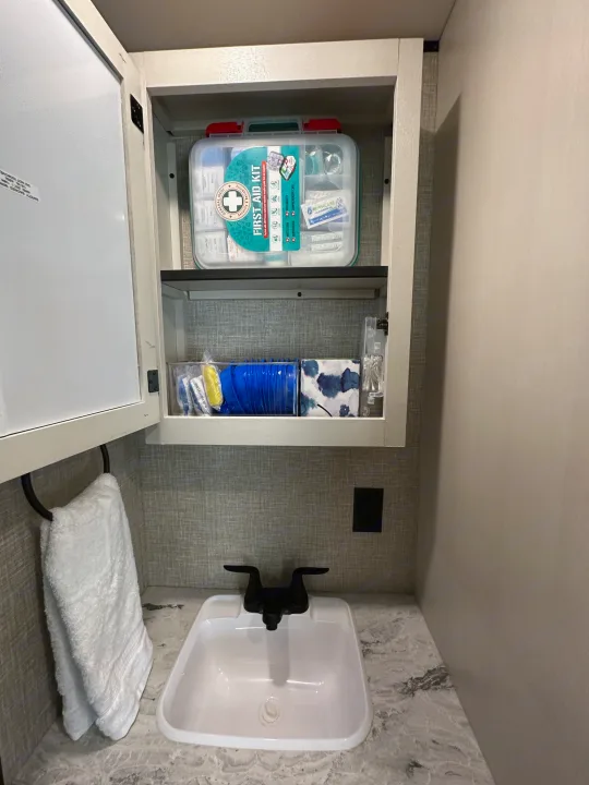 RV rental bathroom's open medicine cabinet reveals a first aid kit and essentials. A sink and towel are below.. Jayco Jay Flight SLX Baja Edition 2023